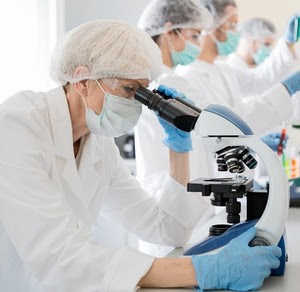 Female scientist in a lab wearing a face mask and looking through a microscope.