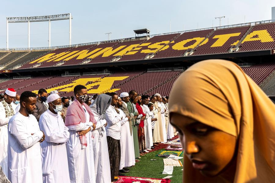Woman in headscarf at the forefront with a line of Muslim men during prayer behind. In the background, sports stadium seating saying 'Minnesota'