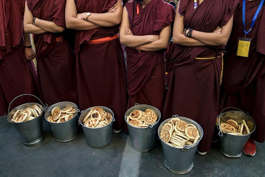 Exile Tibetan Buddhist nuns stand with their arms crossed across their chest. There are buckets of bread at their feet.