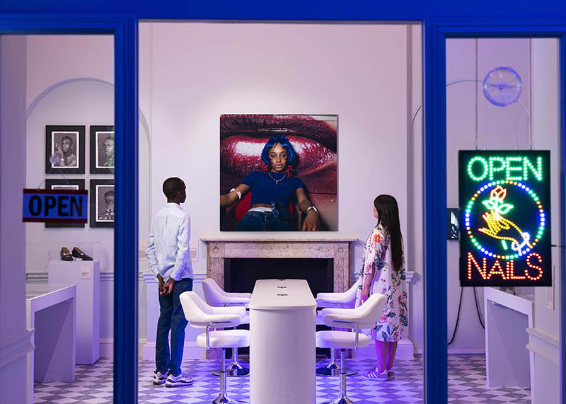 An installation view of The Missing Thread. Two visitors look on at an artwork in a room which has been modelled to resemble a nail salon.