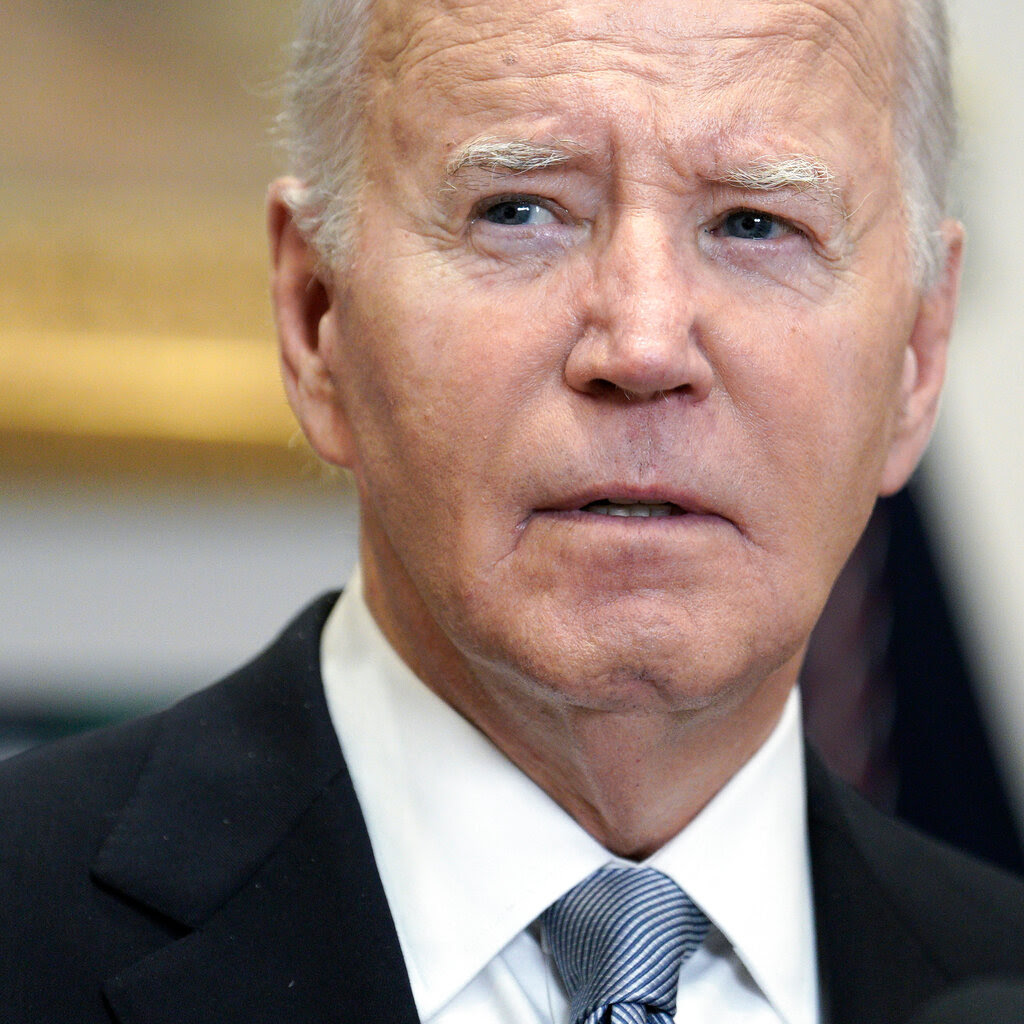 President Biden is speaking, wearing a dark suit and striped tie.