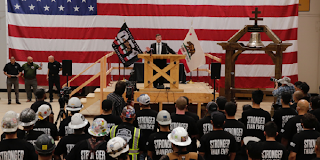 Secretary Walsh speaks on a stage in front of an American flag. Audience members wear shirts reading Stronger than Ever.