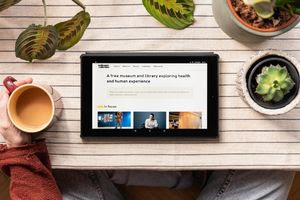 A tablet rests on a wooden table, displaying the home page of the Wellcome Collection website. A person sits in front of the tablet, with their hand resting on a mug on the table. The tablet is surrounded by small plants.