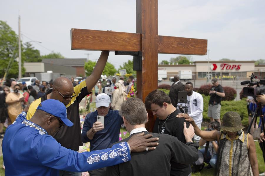 A group of people pray next to a cross at a memorial site for the victims of the Buffalo supermarket shooting.
