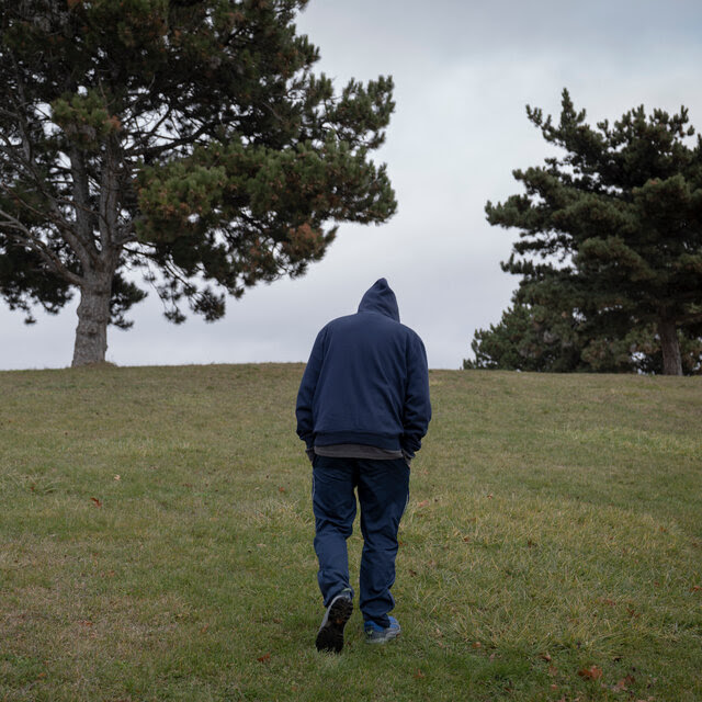 A person wearing a jacket with a hood walks on a grassy field toward a tree.