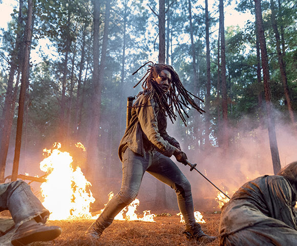 Michonne of The Walking Dead swings her signature blade in a wooded area as a small fire burns in the background.