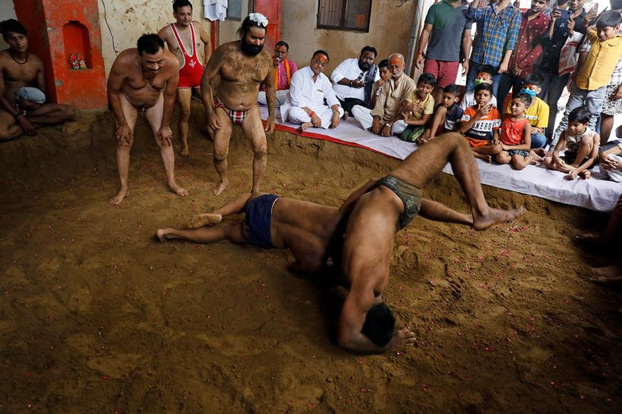 Two Hindu wrestlers grapple on a dirt floor with other wrestlers watching nearby. There are many people watching the match sitting on a cloth and there are some spectators standing behind them.