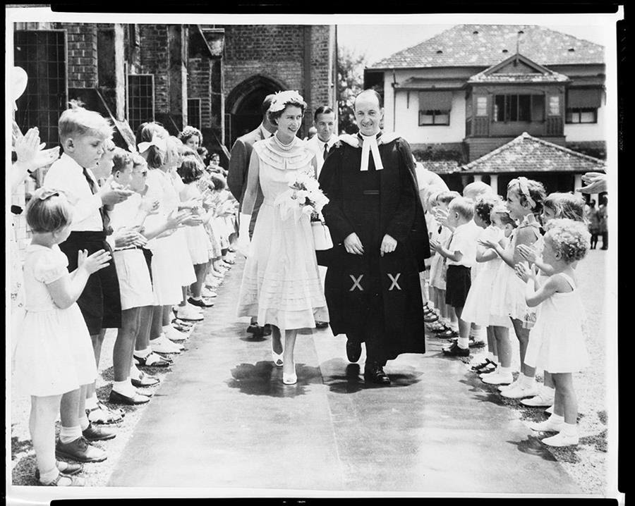 A black and white photo of Queen Elizabeth II leaving St. Michael and All Angels Church in Colombo, Ceylon (now Sri Lanka). There are Sunday school children on either side of the pathway where Queen Elizabeth II is walking.