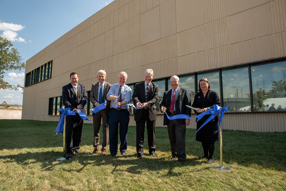 Six people standing in front of a large tan building cutting a blue ribbon.