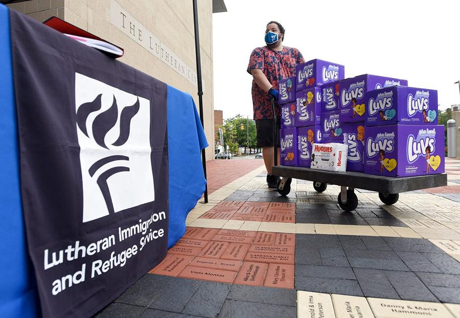 Berny Lopez, an operations specialist for Lutheran Immigration and Refugee, moves a pallet cart of donated diapers. There is a banner for the Lutheran Immigration and Refugee Service in the foreground.