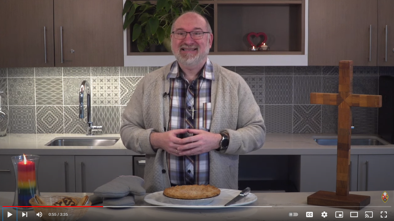 Still from the Moderator's PIE day video. He is standing behind a kitchen counter, a rainbow candle in front of him alongside a pie and a wooden cross.