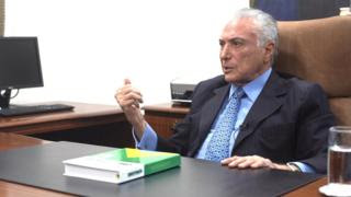 Temer dá entrevista sentado em poltrona e diante de mesa no seu escritório, sobre a qual está um exemplar da Constituição