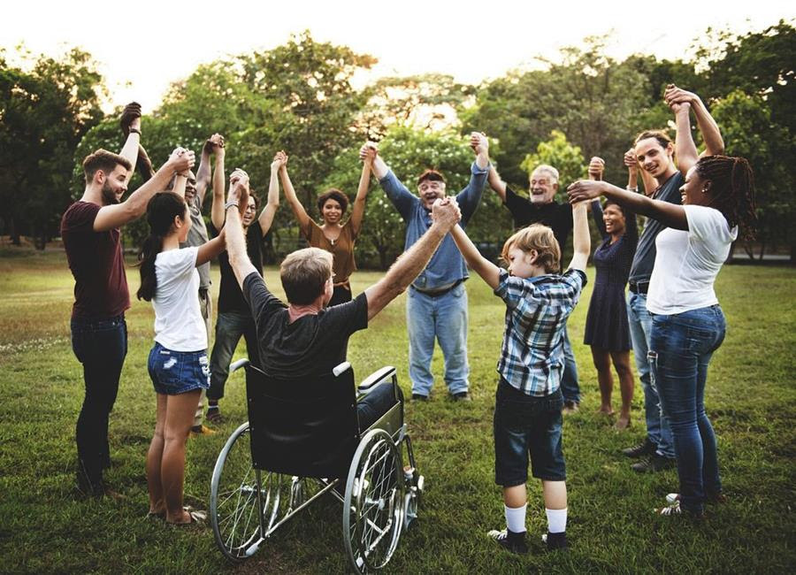 A group of people stand outside in a circle holding hands. One of the people in the circle is in a wheelchair.