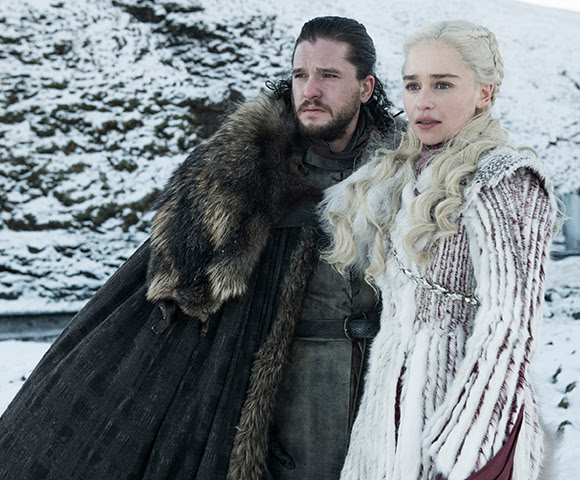 Two Game of Thrones characters dressed in medieval attire watch on in a snowy landscape.