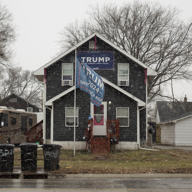 A Trump flag waves outside a gray, shingled house as light snow falls. 