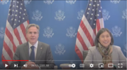 Secretary Blinken and Ambassador Tai smile as they sit at a table in front of a blue backdrop and two U.S. flags. 