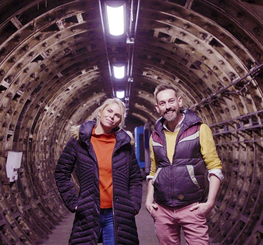 Two smiling people in a circular underground tunnel lined with metal