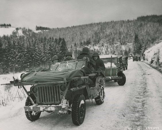 Bullet holes in the windshield of this Jeep are a testament to the faithful service of the Medics of the 84th Division as they evacuate wounded soldiers in their makeshift Jeep ambulance. The Medics are carrying two soldiers wrapped up in blankets to keep them warm in the frigid cold. The litters are strapped to a makeshift frame attached to the flat-hooded Jeep 4x4. This Jeep caravan was part of the 1st Battalion, 334th Infantry Regiment. Image was taken on January 9th, 1945. Thanks to...