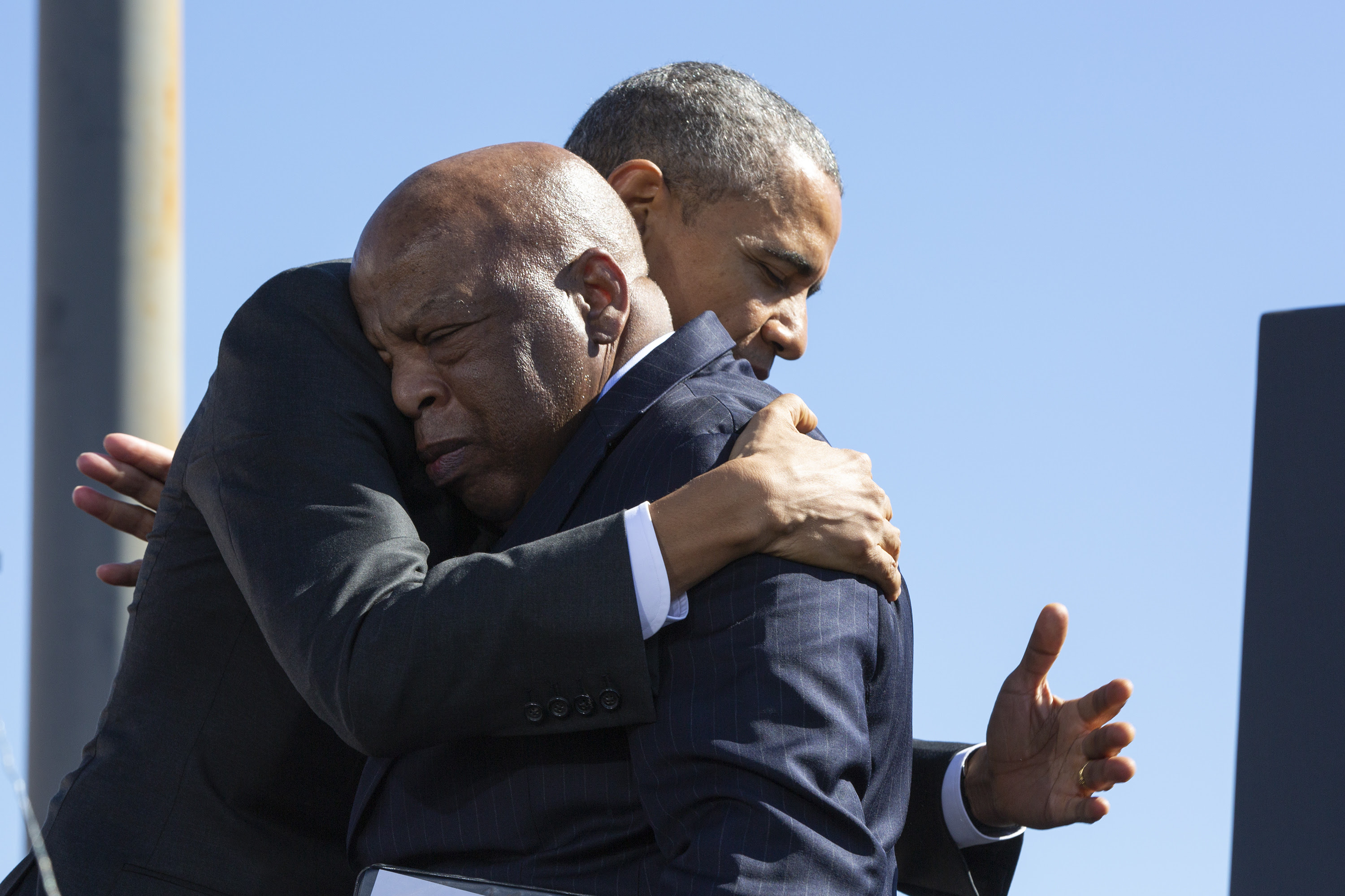 President Obama and Representative John Lewis are shown in profile embracing against a blue sky.