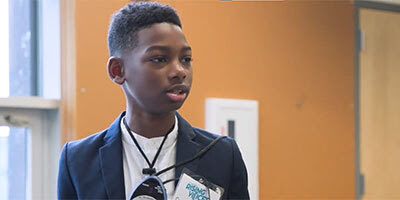 A young Black boy in a suit jacket speaks before a crowd