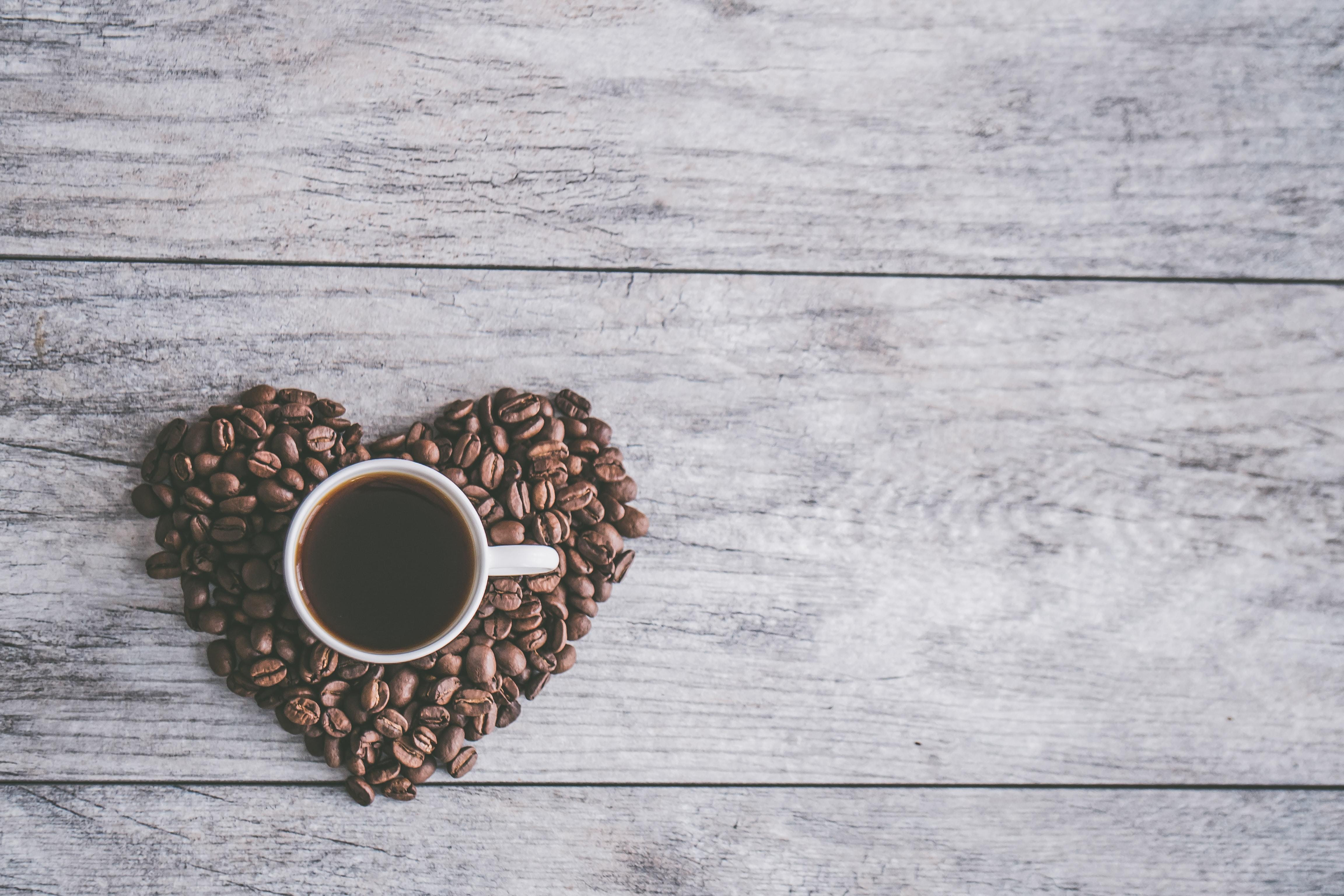 Birds eye photo of a cup of coffee on top of a heart made of coffee beans, on a wooden table.