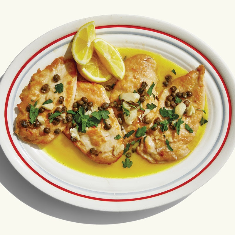 An oval serving plate with four browned chicken cutlets in a garlic-butter sauce, topped with capers, parsley, and lemon wedges.