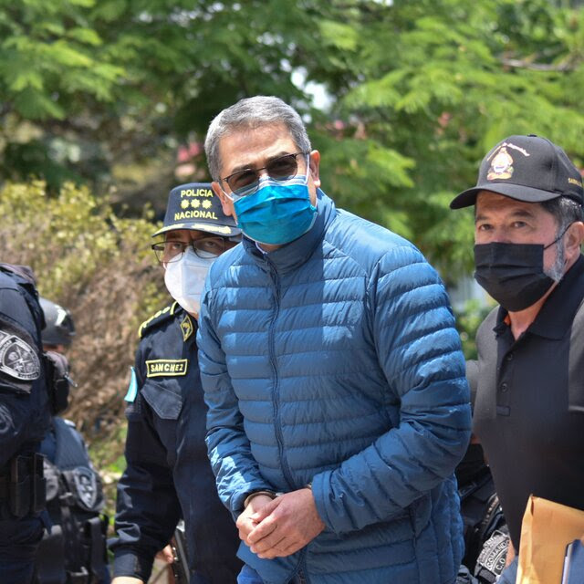 Juan Orlando Hernández, wearing a surgical mask and a blue puffer jacket, is escorted by Honduran police, his hands in handcuffs.