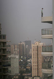 Photo by Feature China / Barcroft Media / Getty Images. Person stands precariously on edge of balcony on very high floor of tall building, city skyline with tall buildings in background.