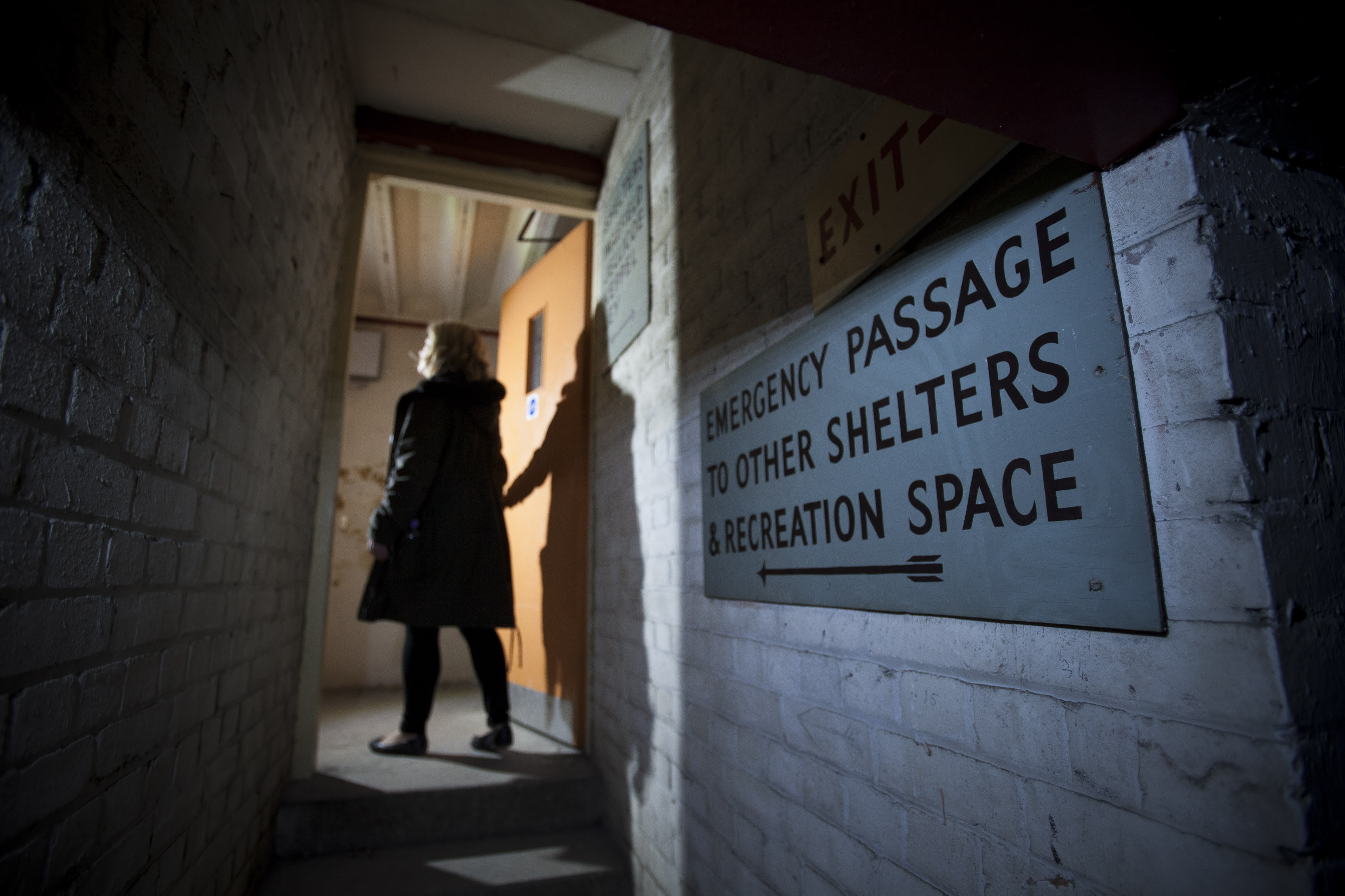 A person and their shadow going through an underground tunnel into a lit up room with a sign on the wall