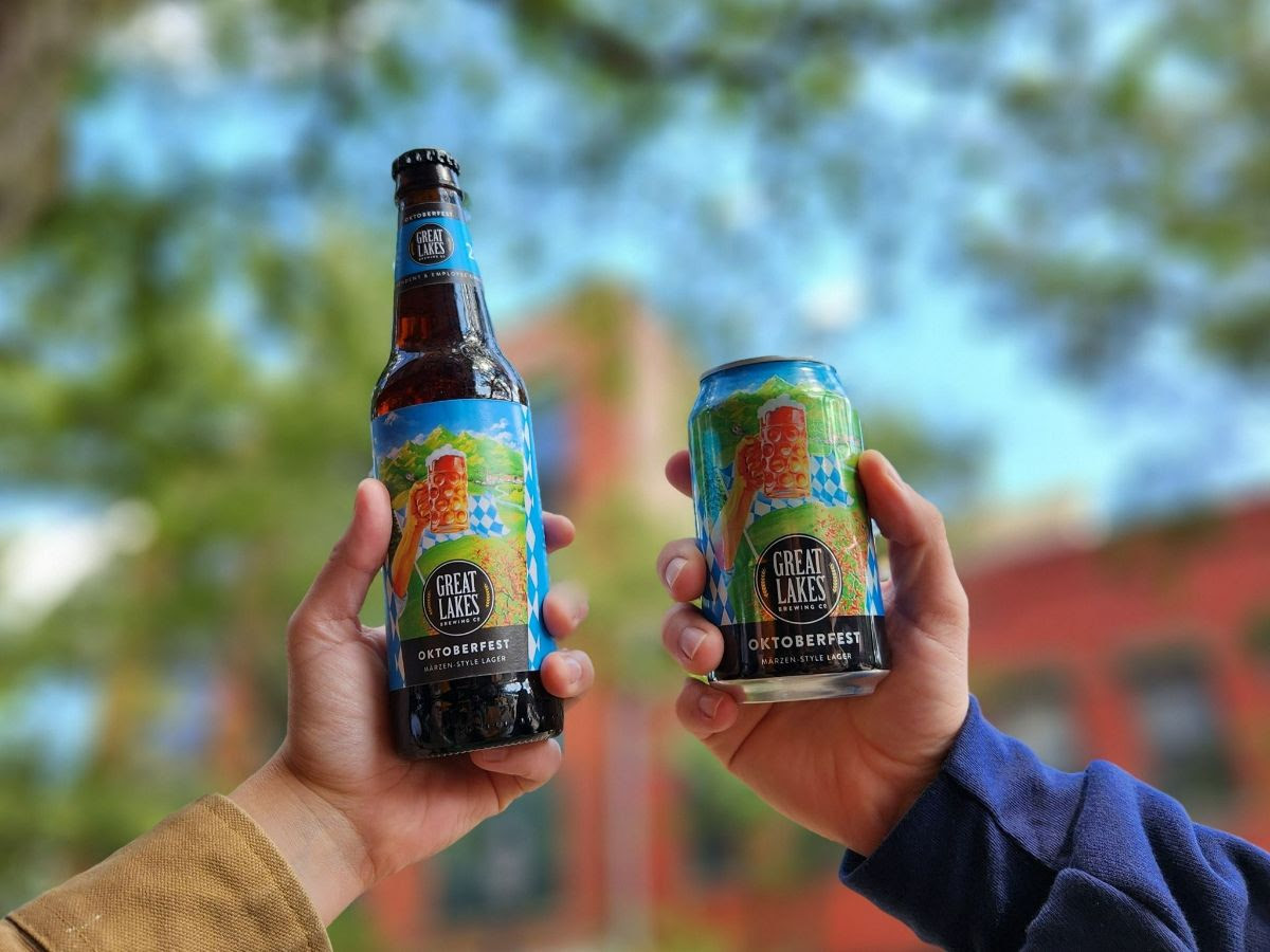 Two people toasting with a bottle and a can of Oktoberfest, with a tree and building in the background.