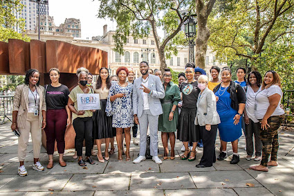 Public Advocate Williams, in a blue plaid suit, stands with all of the speakers at the press conference in City Hall Park. Around twenty women stand with the Public Advocate. 