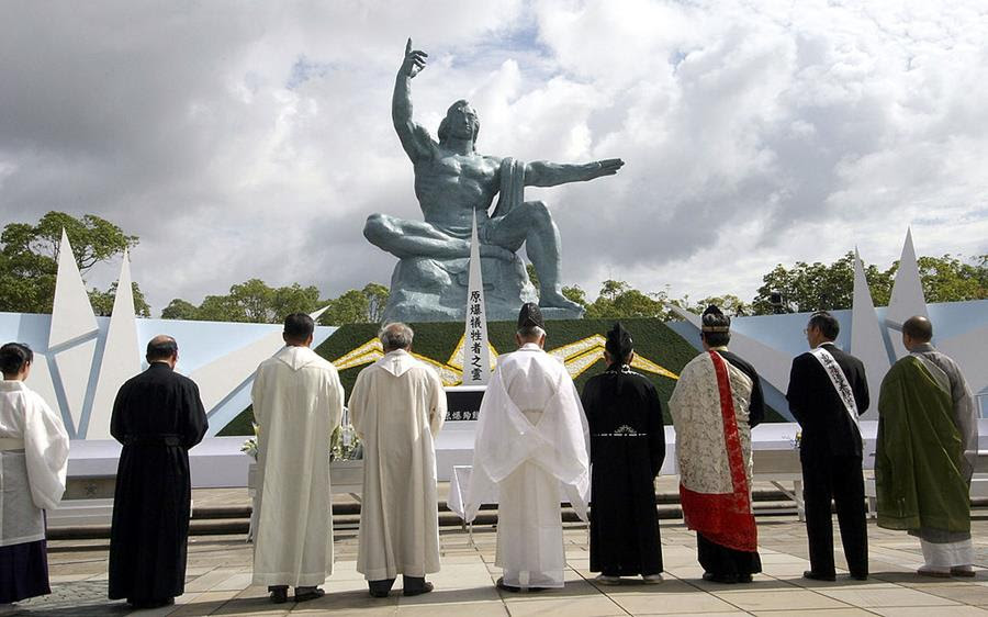 Nine men, many in robes, stand with their backs to the camera in front of a statue of a muscular man pointing to the sky.