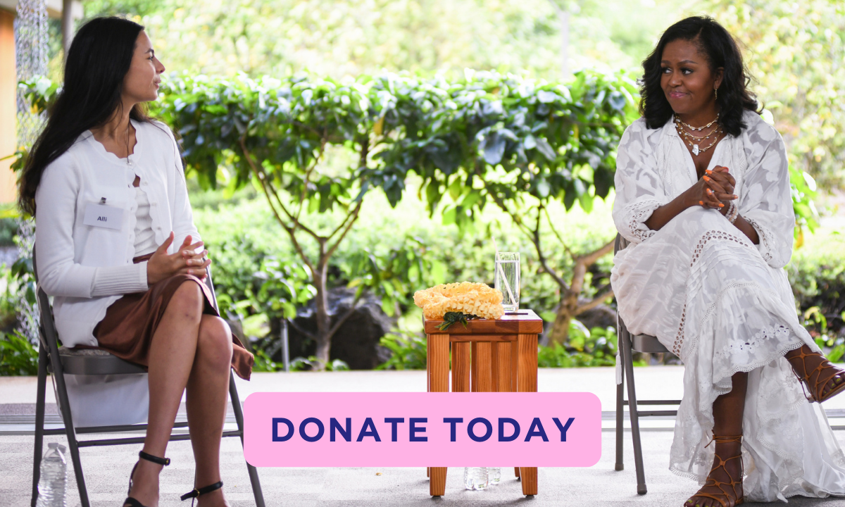 Mrs. Obama sits on the right with her legs crossed, wearing a white patterned dress, looking at a young woman with medium light skin and dark hair, who is wearing a white dress and white cardigan. There is mixed foliage and greenery behind them. A pink button reads “Donate Today” in navy text. 