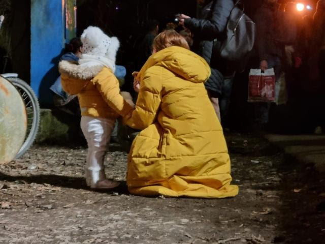 A woman crouches down next to her child to comfort her. It's night time, and they are looking away from the camera.