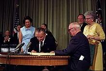 Two men at a desk with a document one is signing with their wives standing behind them