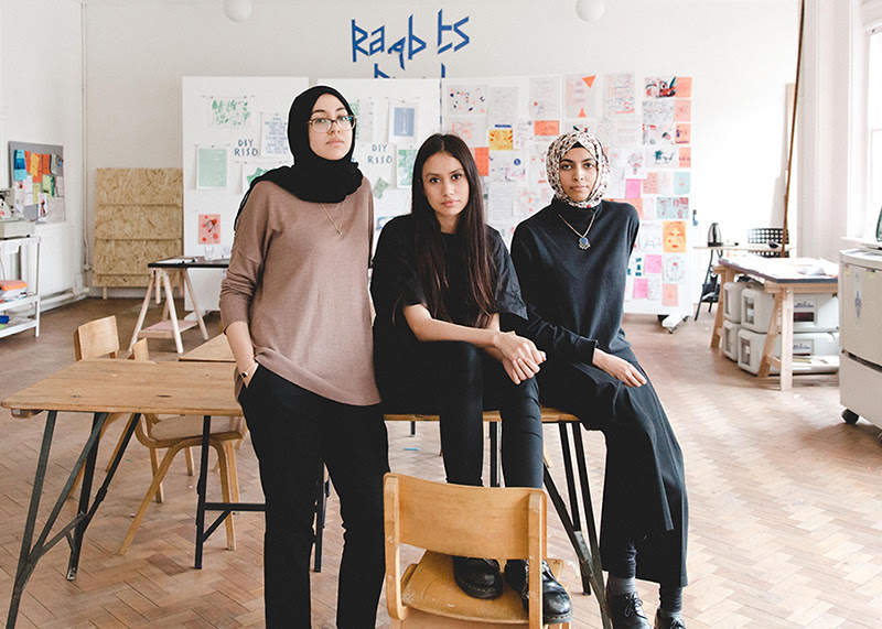 A photo of OOMK in their studio. Three young women sit upon a table. Two wear headscarves and all three are looking straight faced to camera.