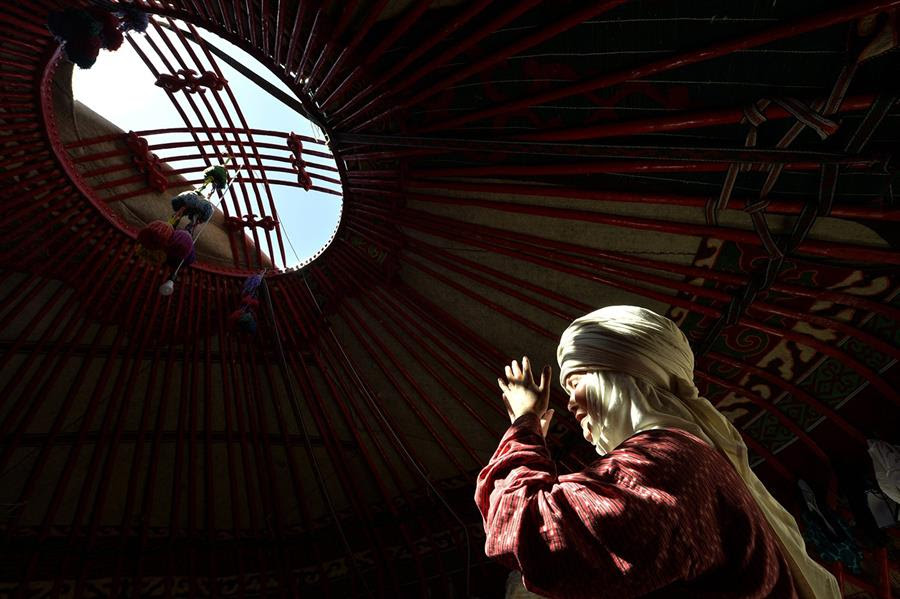 A Kyrgyz woman wearing an elechek, the traditional Kyrgyz headdress for married women, prays inside a yurt. Sunlight is coming through an opening in the ceiling and illuminates the woman.