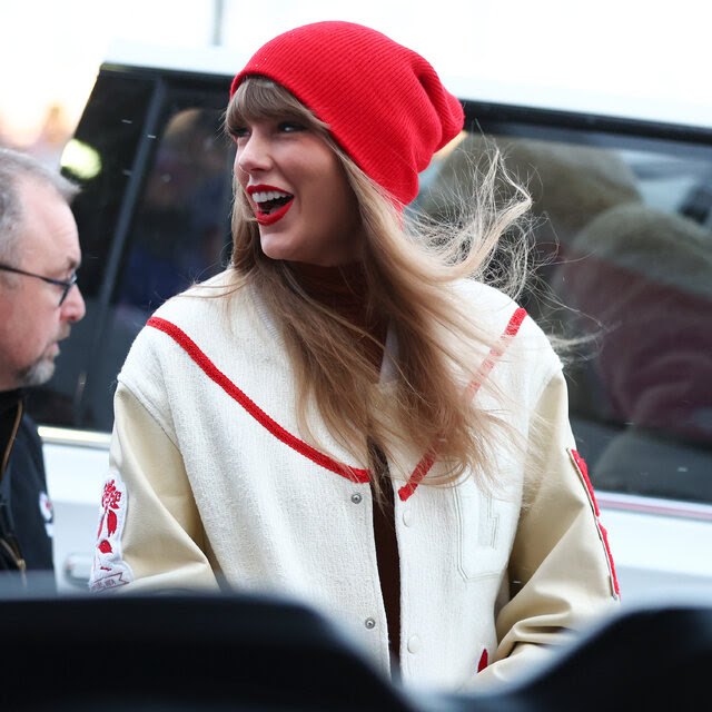 Taylor Swift, in a white jacket and red knit hat, looks to her right and smiles while a man in a black coat walks behind her.