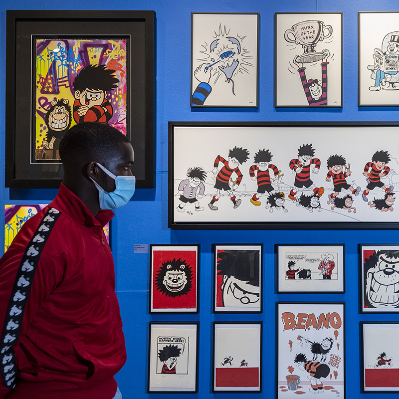 A view of the Beano exhibition shop. There are lots of Beano related prints on the wall. A young Black man wearing a red jacket is looking at the prints.