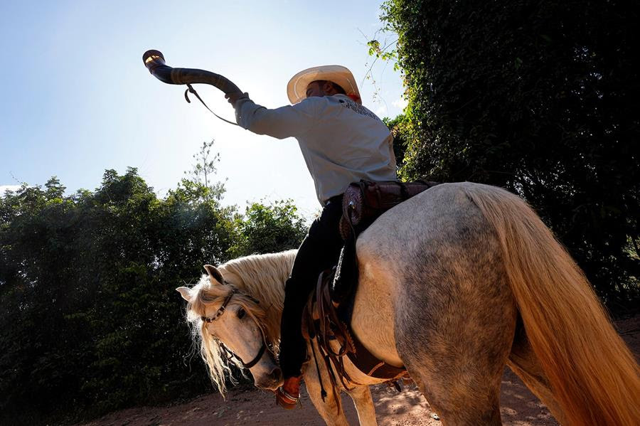 A man on horseback plays a Berrante, an ox horn musical instrument. He is participating in a parade that culminates the religious tradition, "Folia do Divino Espirito Santo" or Feast of the Divine.