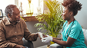 A woman wearing gloves, scrubs and a stethoscope holds the hand of an older man.