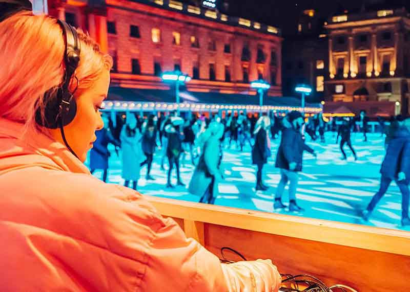 A woman is DJing. In front of her you can see an ice rink, lit blue, with people skating around.