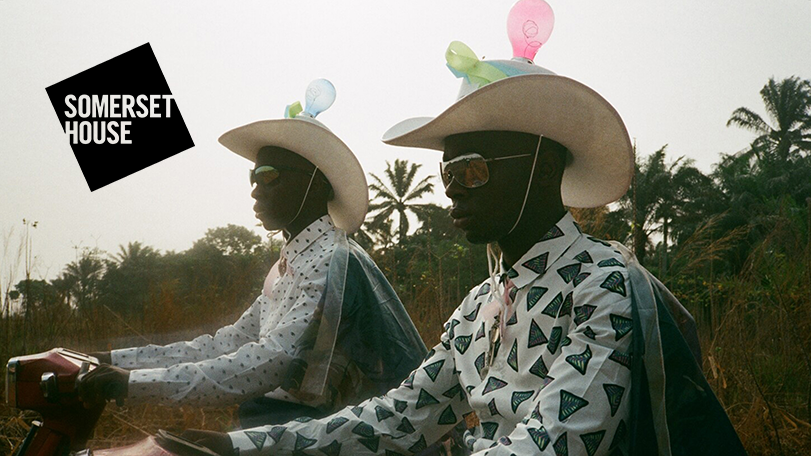 A still from a film by Akinola Davies Jr. It shows two Black men wearing pink-white cowboy hats, sunglasses and buttoned up shirts, riding scooters.