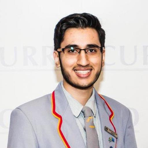 Headshot του Waheed Amanjee, ο οποίος χαμογελά και κρατάει μια σχολική στολή που περιλαμβάνει πουκάμισο και γκρι γραβάτα και blazer