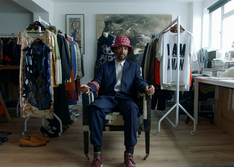 A still from The Missing Thread short documentary. Harris, one of the curators of the exhibition, sits on a chair in a studio space with two racks of clothing behind him. He wears a nicely tailored navy suit and a red and blue checked hat.