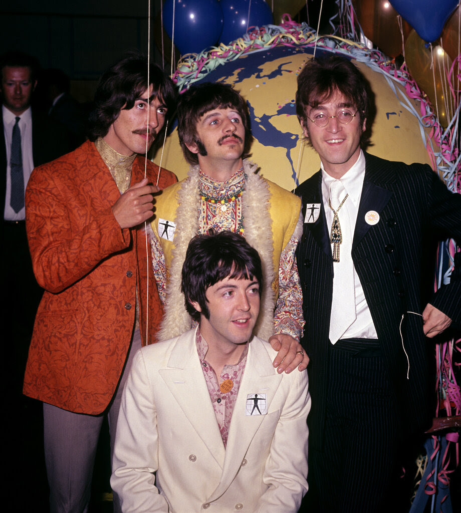 The Beatles, with shaggy hair and mustaches, stand in front of a backdrop that looks like Earth surrounded by streamers and balloons.