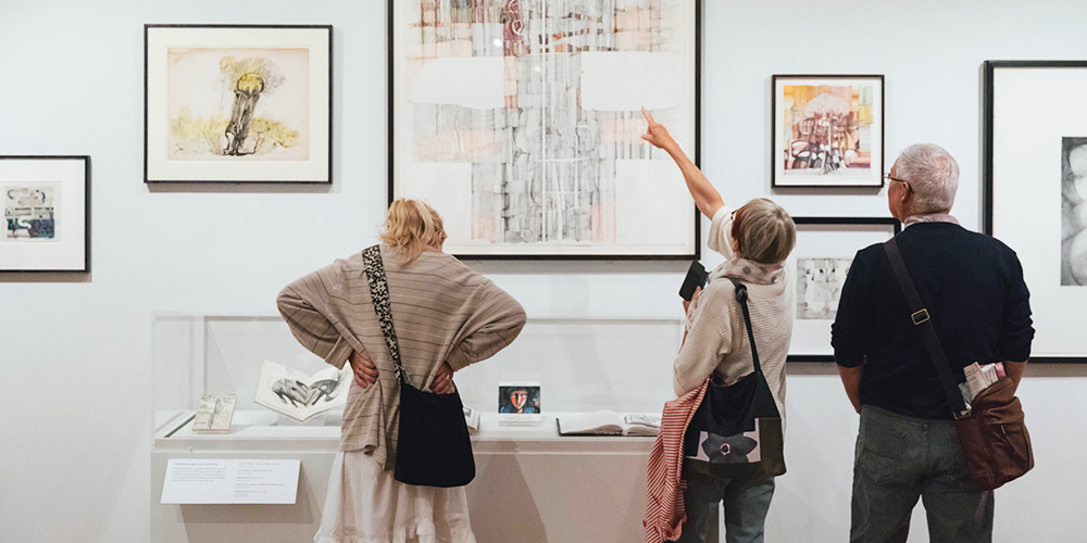 Three figures with their backs to us, looking at art on a gallery wall and in a glass case