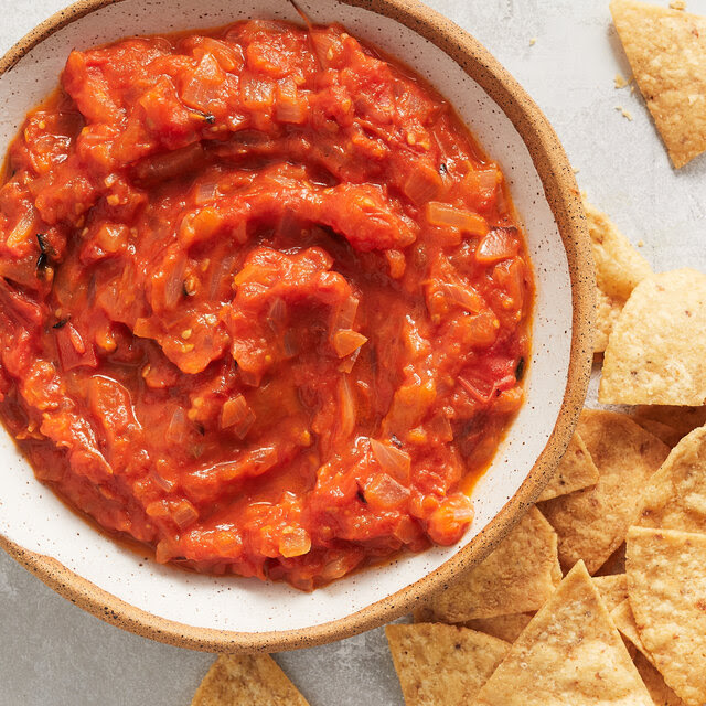 A ceramic bowl holds a red-orange chunky salsa. On a marble surface beneath it sits a handful of tortilla chips.