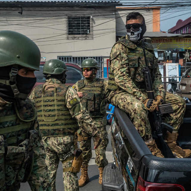 Several soldiers, some with their faces covered, stand on a city street, while one sits in the bed of a pickup truck. 