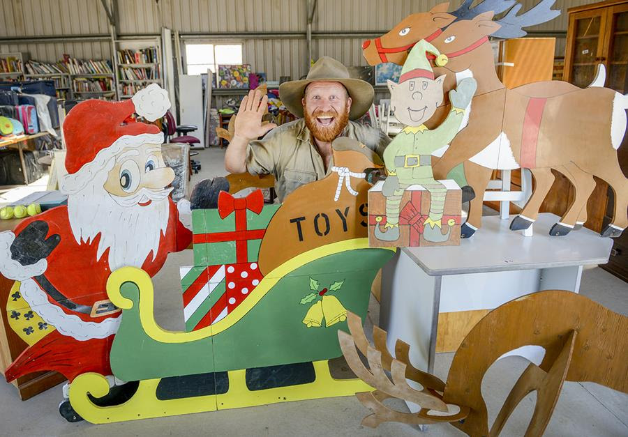 Waste Warrior Wombat waving and standing behind a wooden santa and sleigh with reindeer and presents at a buyback centre.
